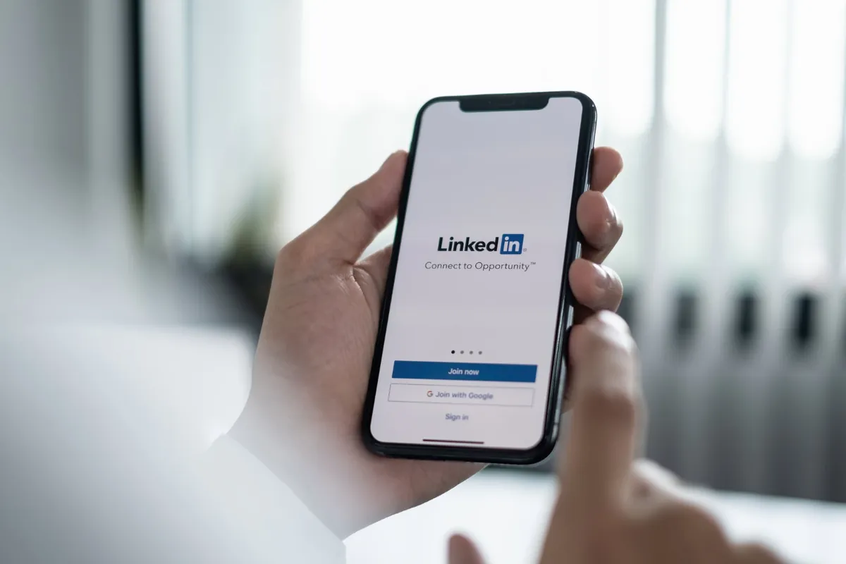 Mão segurando um smartphone com a tela de cadastro do LinkedIn para empresas exibindo os botões "Join now" e "Sign in"
